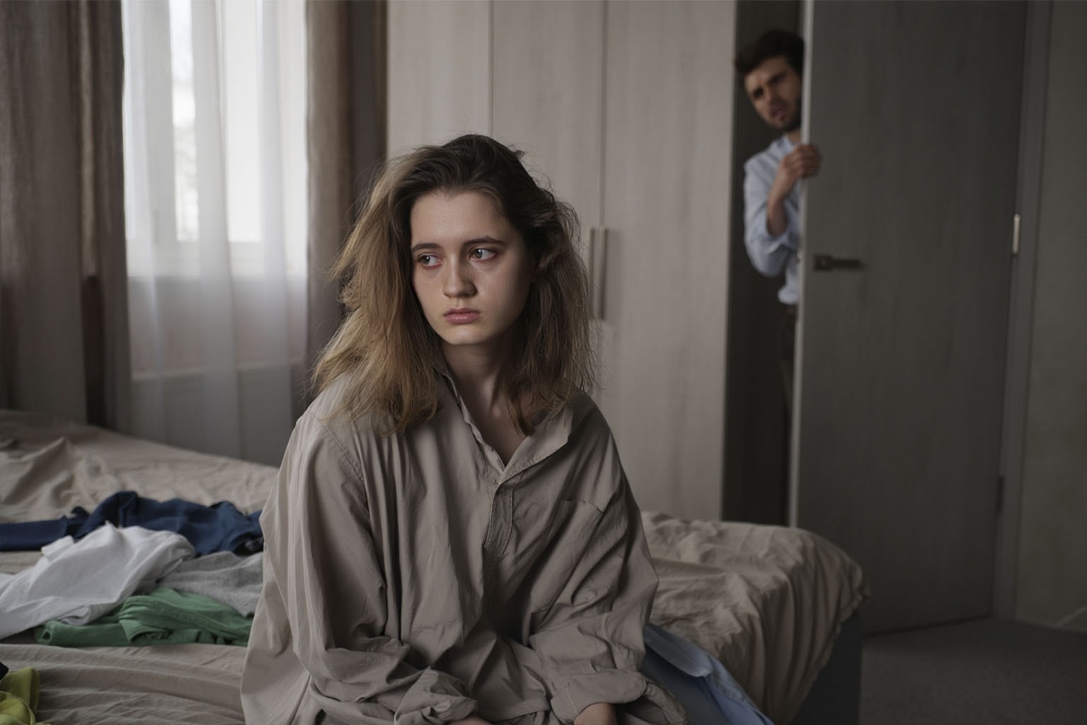 Женщина грустная и неопрятная сидит на кровати, муж переживает